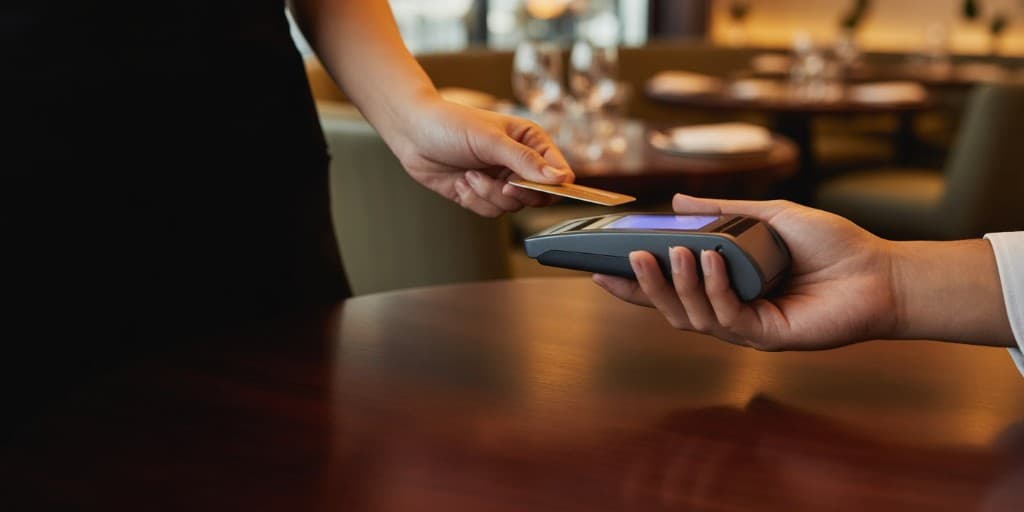 Επαφή πληρωμής: χέρι πελάτη με κάρτα πάνω σε τερματικό POS, θερμό φωτισμό, θολό εστιατόριο στο φόντο